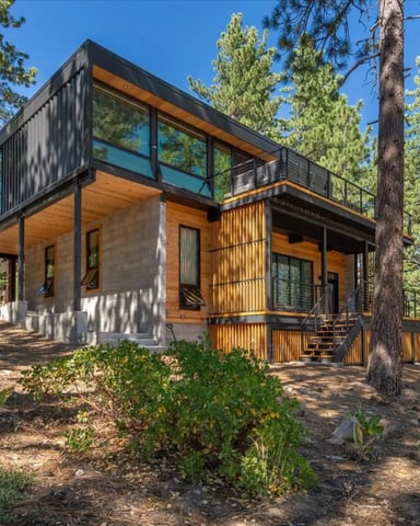 Modern cabin with black metal frame, wood siding, and large windows nestled among tall pine trees in forest setting.