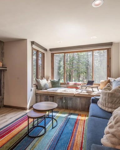 Modern living room with fireplace, blue sofa, window seat overlooking forest, and colorful striped rug