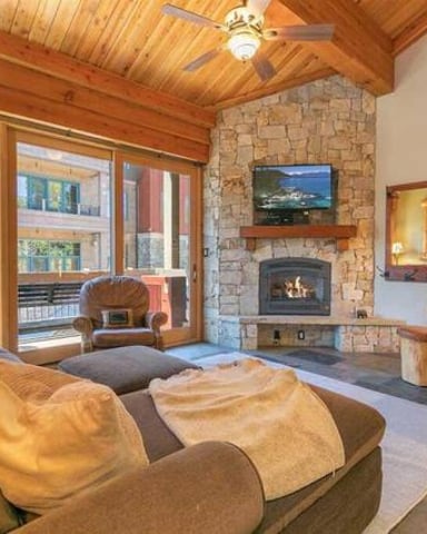 Spacious living room with wood beams, stone fireplace, gas stove, comfortable seating, and glass doors opening to a deck