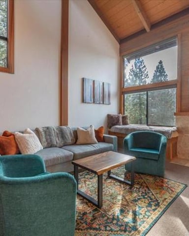 Modern living room with wood-paneled ceiling, gray sofa, teal armchairs, fireplace, and forest views through large windows