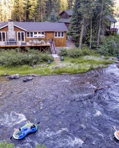 Riverside cabin with wooden deck surrounded by tall evergreen trees, two people tubing on river in foreground