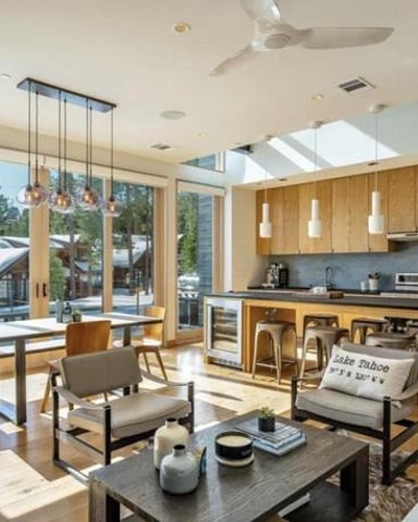 Modern open-plan kitchen and living room with wood cabinetry, stainless steel appliances, dining table, and large windows overlooking forest and mountains.