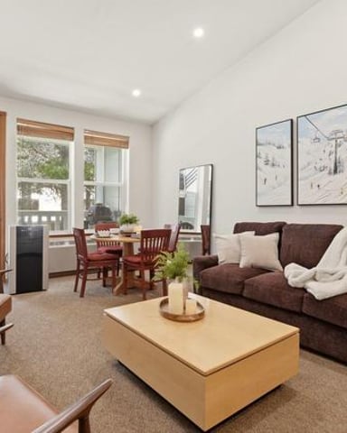 Bright living room with brown sofa, wooden furniture, dining table, and glass doors opening to a deck with forest view
