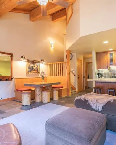 Open-plan living room with stone fireplace, exposed wood beams, gray seating, orange kitchen bar, and wood cabinets