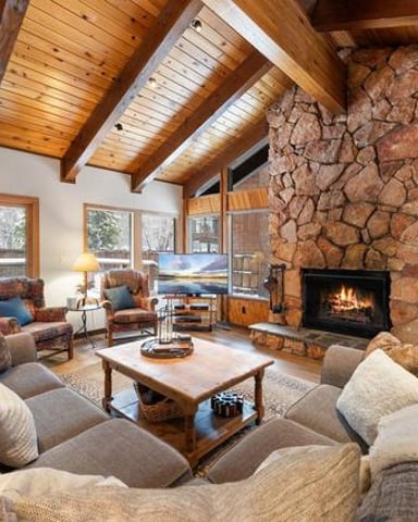 Spacious living room with wood beams, stone fireplace, upholstered seating, and large windows overlooking forest