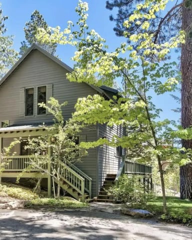 Modern gray two-story cabin nestled among tall pine trees with front deck and stairs