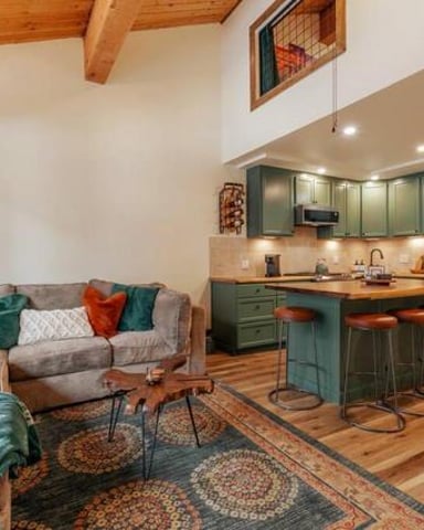 Open-plan living room and kitchen with exposed wood beams, gray sofa with colorful pillows, green cabinetry, and wooden bar seating