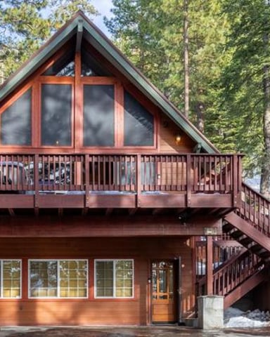 A-frame cabin with reddish-brown wood siding, large upper windows, and wraparound deck surrounded by tall pine trees and snow.
