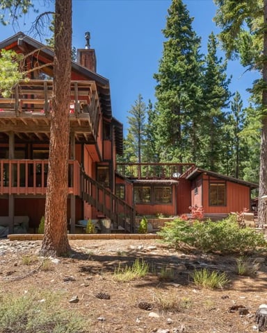 Red cabin with elevated deck and chimney surrounded by tall pine trees and outdoor fireplace