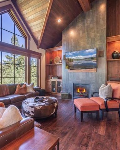 Spacious living room with leather sectional sofa, fireplace, wood beams, large windows overlooking trees, and built-in shelving.