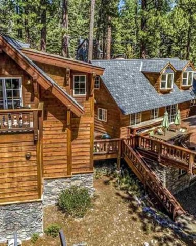 Wooden cabin with stone foundation nestled among tall pine trees, featuring multiple decks and a small creek running beneath