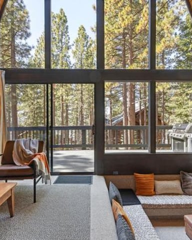 Modern cabin bedroom with wood walls, large windows overlooking forest, bed, seating area, and deck access