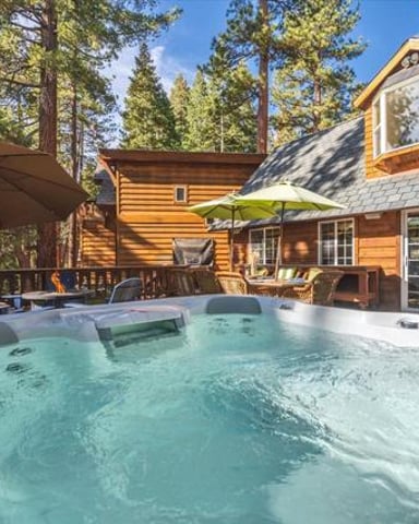 Hot tub on deck of log cabin surrounded by tall pine trees with clear blue sky