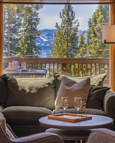 Living room with gray sofa, armchairs, and round table overlooking mountain lake and pine forest from balcony