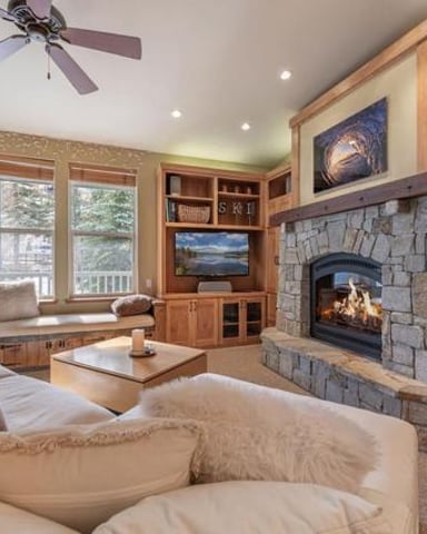 Spacious bedroom with fireplace, wood cabinetry, ceiling fan, and large windows overlooking trees