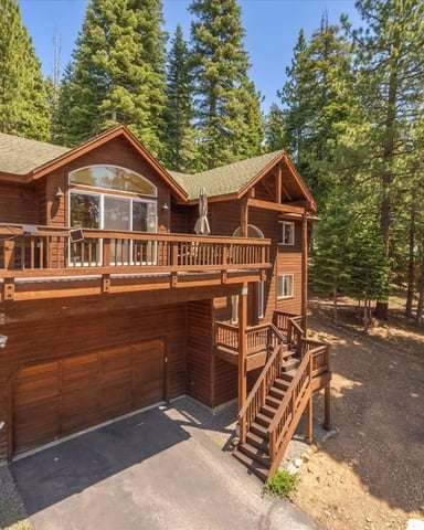 Two-story wood cabin with peaked roof and wraparound deck surrounded by tall pine trees in forest setting.
