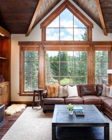 Modern living room with vaulted wood ceiling, large windows, brown leather sectional, fireplace, and forest views