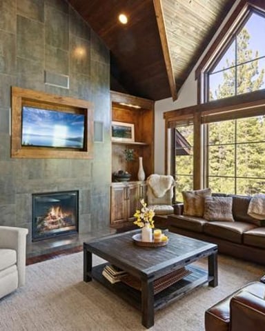 Modern living room with fireplace, leather seating, large windows overlooking forest, and vaulted ceiling with skylights