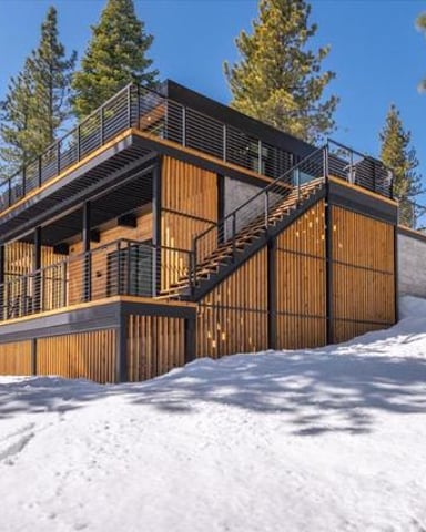 Modern wooden and metal cabin with multiple decks surrounded by tall pine trees and snow
