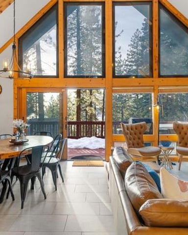 Modern cabin interior with vaulted wood ceilings, large windows overlooking forest, leather seating, wood stove, and dining area.