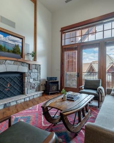 Living room with stone fireplace, wood mantel, framed mountain landscape, large windows with mountain views, and upholstered seating around coffee table