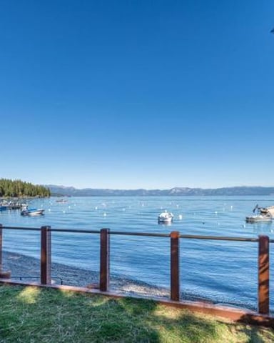 Wooden dock railing overlooking calm blue lake with moored boats, forested shoreline, and distant mountains under clear sky