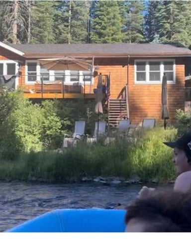 Two people in river looking at orange and white cabin with stone chimney surrounded by tall evergreen trees