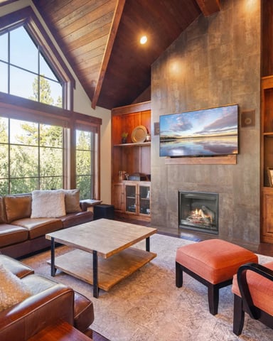 Modern living room with vaulted wood ceiling, leather seating, gas fireplace, built-in wood cabinetry, and large windows overlooking forest.