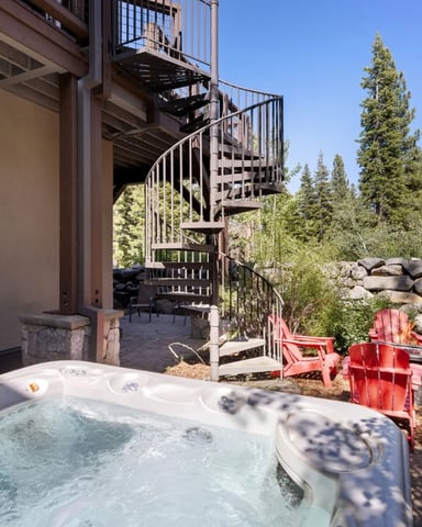 Hot tub on patio with red Adirondack chairs, spiral staircase, stone wall, and pine forest backdrop