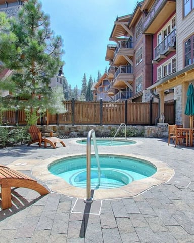 Resort courtyard with hot tubs, wooden lounge chairs, multi-story lodge buildings, and pine trees under clear sky