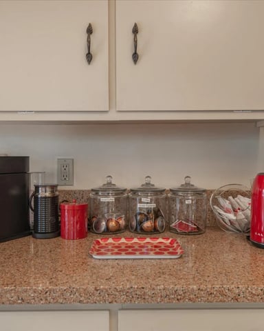 Kitchen counter with granite countertop, red kettle, coffee maker, storage containers, and white cabinetry above