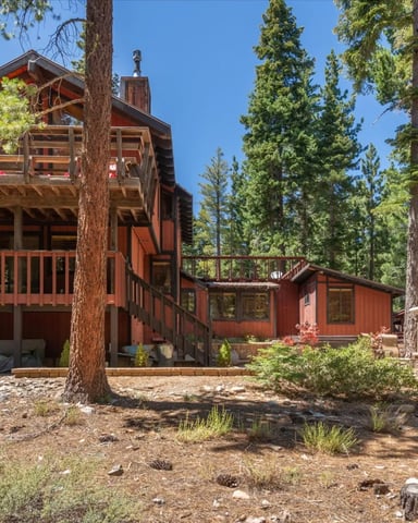 Red-brown wooden cabin with elevated deck and chimney surrounded by tall pine trees and outdoor fireplace