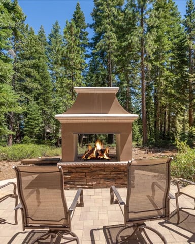 Outdoor patio with metal chairs surrounding a stone fireplace with active fire, surrounded by tall pine trees