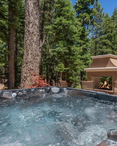 Hot tub surrounded by tall pine trees with outdoor fireplace and seating area in forest clearing
