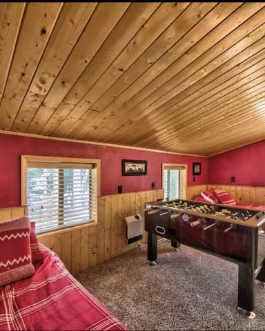 Recreation room with red walls, wood paneling, foosball table, bed, and windows overlooking forest.