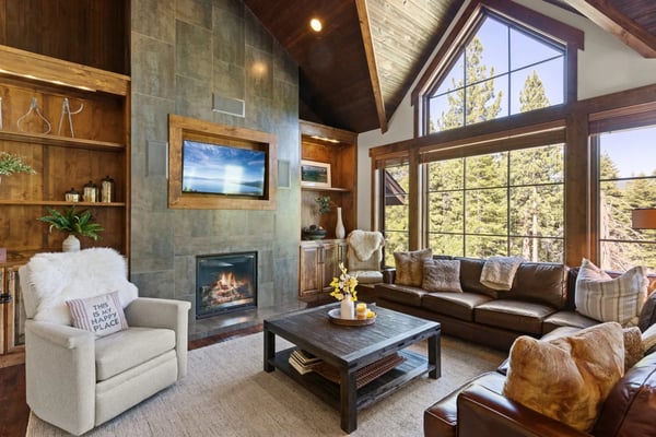 Modern living room with stone fireplace, wood paneling, leather sofa, and large windows overlooking pine forest