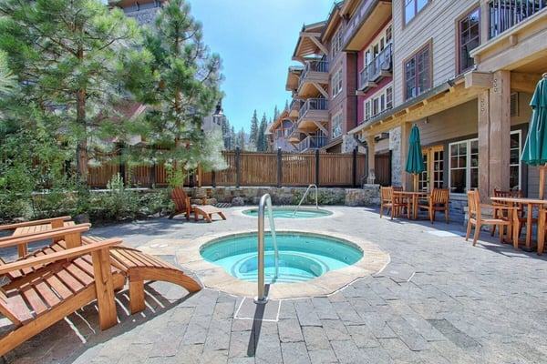 Resort courtyard with two hot tubs, wooden lounge chairs, multi-story lodge building, and pine trees under clear sky