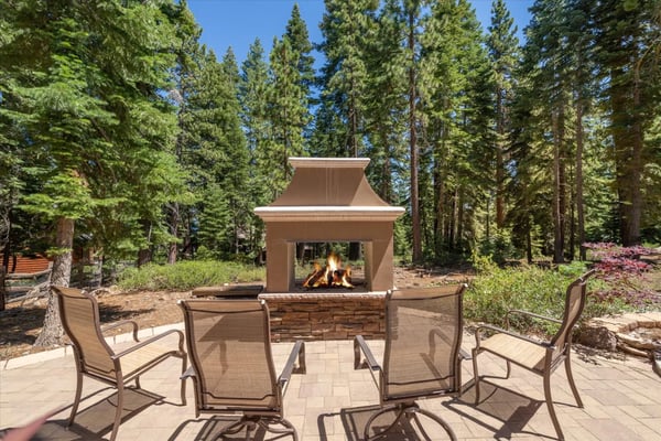Outdoor patio with metal chairs surrounding a stone fireplace with active fire, surrounded by tall pine trees