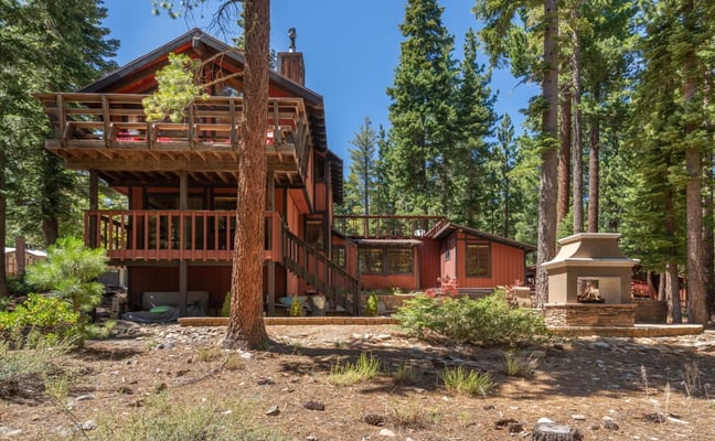 Red-brown wooden cabin with elevated deck and chimney surrounded by tall pine trees and outdoor fireplace