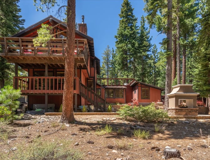 Red-brown wooden cabin with multi-level decks nestled among tall pine trees and clear blue sky